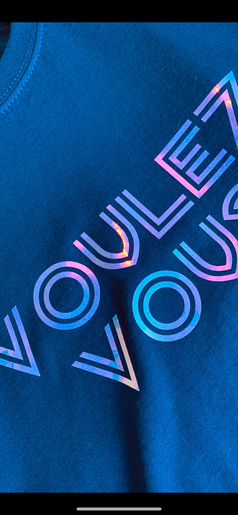 May include: Navy blue t-shirt with the word "VOUS" in a holographic, iridescent design. The text is angled across the shirt, creating a modern and eye-catching look. The shirt is made of a soft fabric.