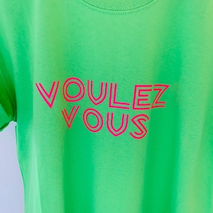 May include: A neon green t-shirt with the words "VOULEZ VOUS" in bright pink, outlined in a darker pink. The text is centered on the shirt, which has a crew neck and short sleeves. The shirt is made of a soft material.