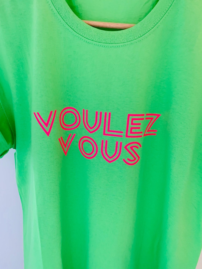 May include: A neon green t-shirt with the words "VOULEZ VOUS" in bright pink, outlined in a darker pink. The text is centered on the shirt, which has a crew neck and short sleeves. The shirt is made of a soft material.