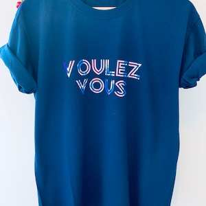 May include: A navy blue t-shirt with rolled-up sleeves. The shirt features the text "VOULEZ VOUS" in a colorful, stylized font. The letters are outlined in white with blue and pink accents.