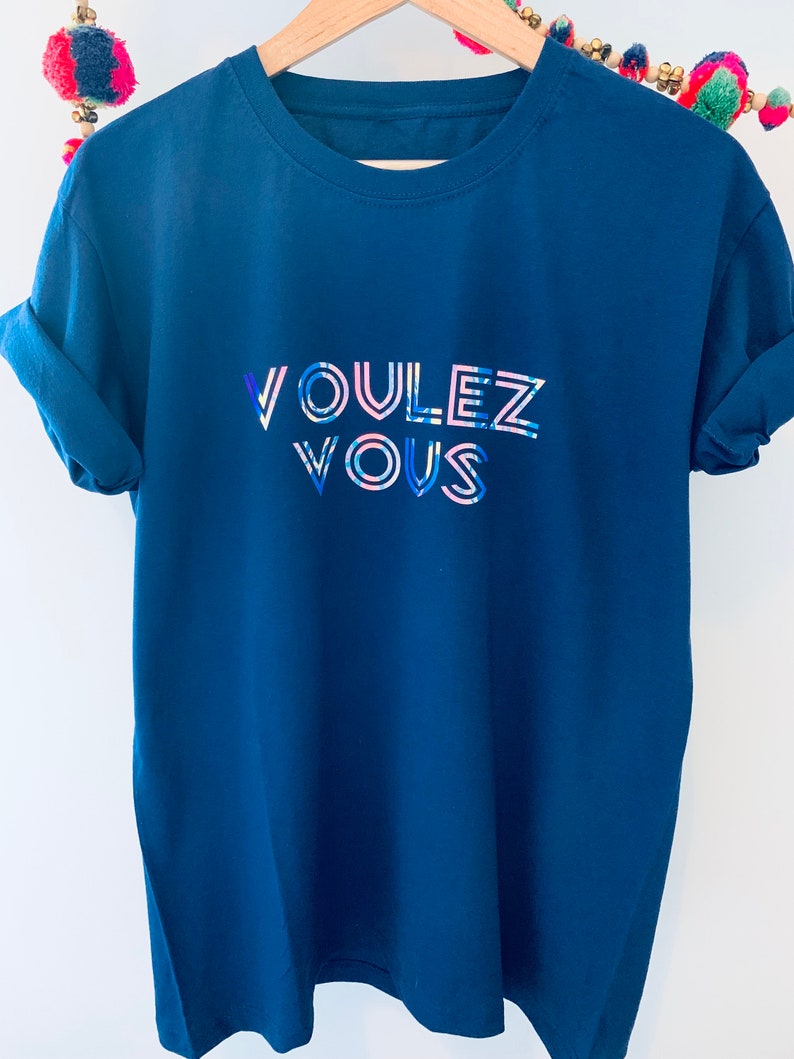 May include: A navy blue t-shirt with rolled-up sleeves. The shirt features the text "VOULEZ VOUS" in a colorful, stylized font. The letters are outlined in white with blue and pink accents.