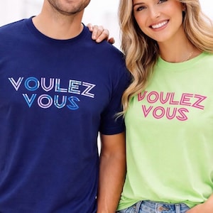 May include: Two people wearing matching t-shirts with the text "Voulez Vous" in a colorful font. One shirt is navy blue, the other is lime green. Both are smiling and wearing casual clothing.