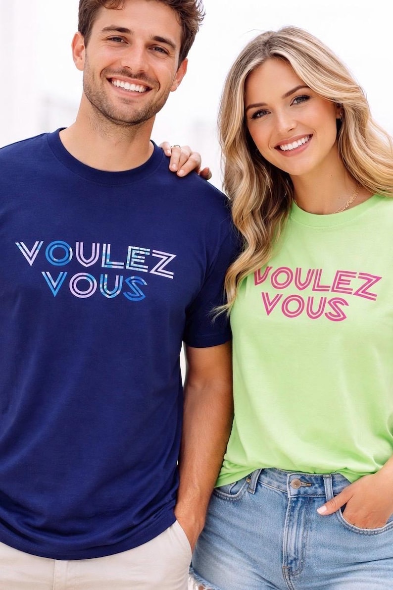 May include: Two people wearing matching t-shirts with the text "Voulez Vous" in a colorful font. One shirt is navy blue, the other is lime green. Both are smiling and wearing casual clothing.