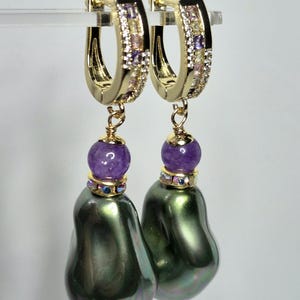 Puede incluir: Pendientes de aro dorados con un diseño colgante. Cada pendiente presenta una gran perla barroca verde oscuro, una cuenta morada y un aro dorado adornado con pequeñas piedras preciosas de colores. Los pendientes se muestran sobre un fondo blanco.