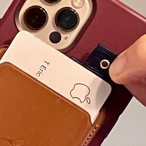 Puede incluir: Una funda de tel&eacute;fono de cuero marr&oacute;n con un porta tarjetas blanco adjunto. El porta tarjetas tiene el logotipo de Apple y el texto "T. Eric" impreso en &eacute;l.