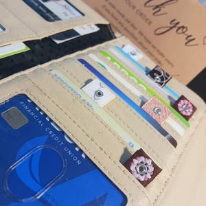 Puede incluir: Una cartera de cuero beige con m&uacute;ltiples tarjetas de cr&eacute;dito y tarjetas de regalo en su interior. La cartera tiene un cierre de cremallera y un adorno floral decorativo.