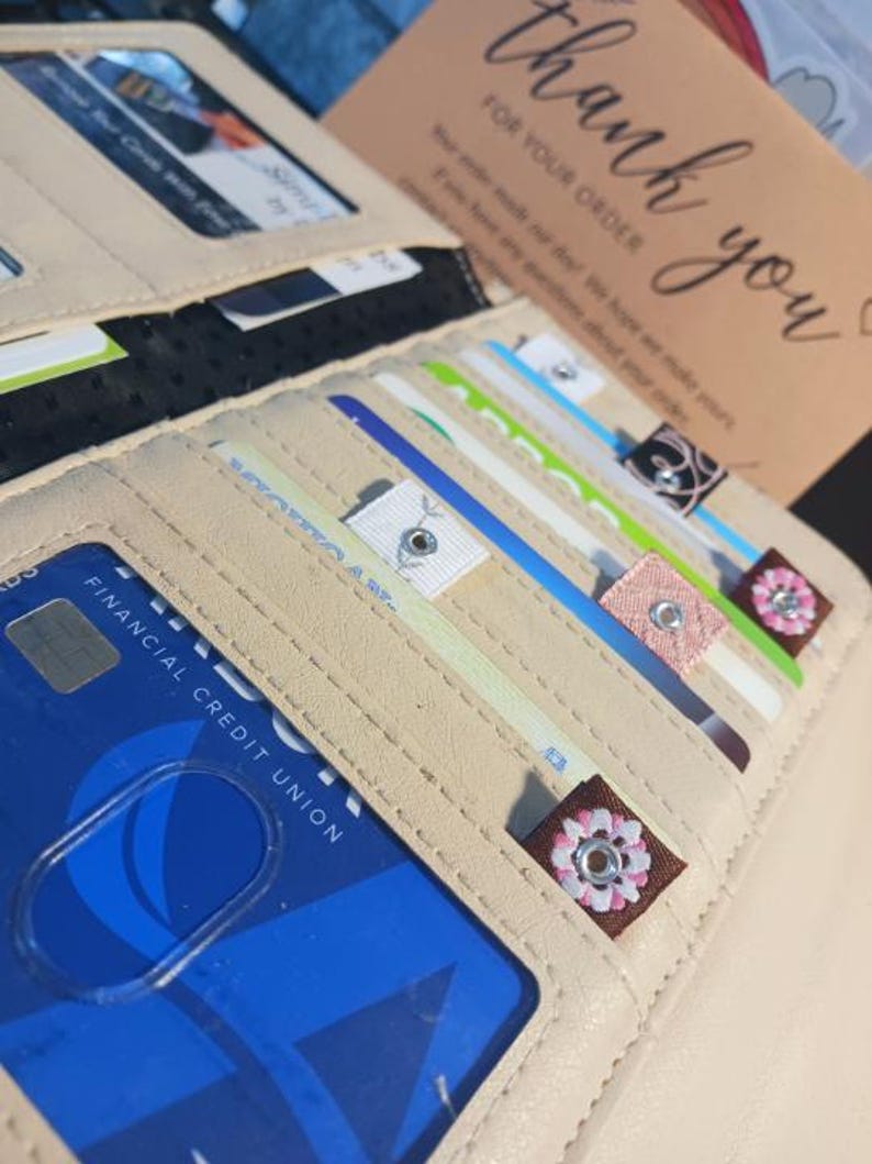 Puede incluir: Una cartera de cuero beige con m&uacute;ltiples tarjetas de cr&eacute;dito y tarjetas de regalo en su interior. La cartera tiene un cierre de cremallera y un adorno floral decorativo.