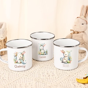以下が含まれることがあります： 銀色の縁と持ち手が付いた、白いエナメルマグカップ3つ。各マグカップには、ウサギと文字と名前（Quincy、Lucien、Julius）の水彩イラストが描かれています。マグカップはイースターをテーマにした要素で飾られています。