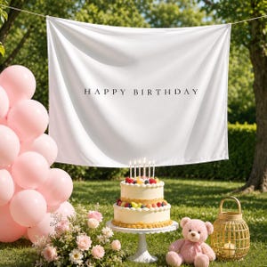 May include: A white fabric backdrop with the black text HAPPY BIRTHDAY, a two-tier cake topped with fruit and candles, a cluster of light pink balloons, a bouquet of pink and white flowers, and a light brown teddy bear with a lantern.