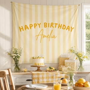 Può includere: Scena di festa di compleanno con uno sfondo a righe gialle e bianche con la scritta "HAPPY BIRTHDAY Amelia". Una torta con fette di limone è su un supporto di legno. Fiori, regali e limoni decorano il tavolo.