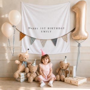 May include: A white fabric backdrop with the text Happy 1ST BIRTHDAY EMILY, featuring a bunting banner, two cream balloons, a large gold number one balloon, three teddy bears wearing party hats, and a child in a light pink dress.