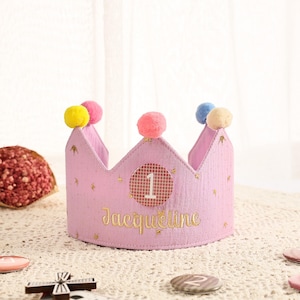 May include: A pink fabric crown with the number "1" on a circular sticker and the name "Jacqueline" embroidered in gold. The crown is decorated with colourful pom-poms and gold star accents, suitable for a first birthday celebration.