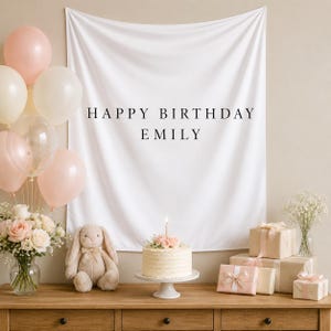 May include: A white fabric backdrop featuring the black text HAPPY BIRTHDAY EMILY, displayed with a cluster of pale pink and cream balloons, a frosted cake with a single candle, a plush rabbit toy, and several gift boxes wrapped in paper.