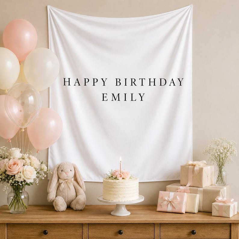 May include: A white fabric backdrop featuring the black text HAPPY BIRTHDAY EMILY, displayed with a cluster of pale pink and cream balloons, a frosted cake with a single candle, a plush rabbit toy, and several gift boxes wrapped in paper.