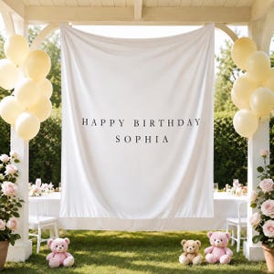 May include: A white fabric backdrop featuring the black text HAPPY BIRTHDAY SOPHIA, flanked by clusters of pale yellow balloons and pink teddy bears, set within a white wooden structure adorned with pink rose bushes on both sides.