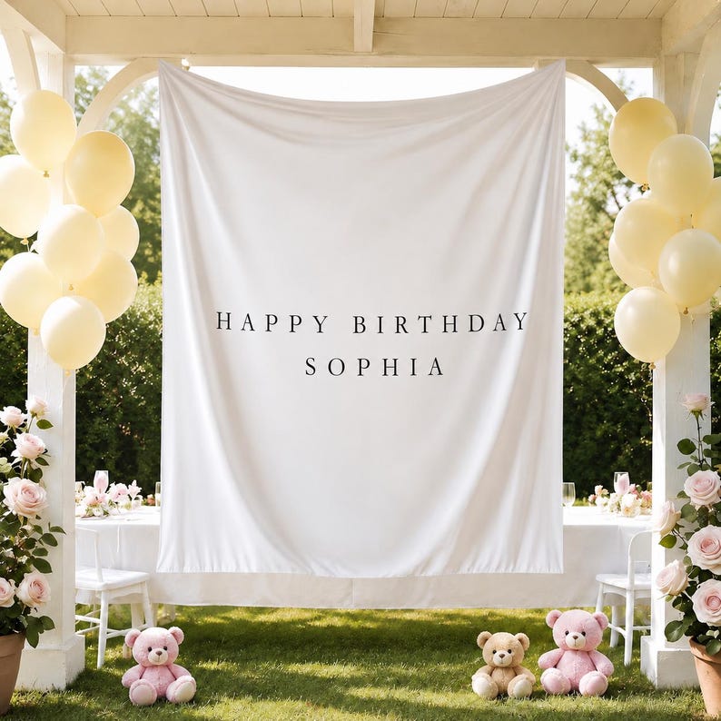 May include: A white fabric backdrop featuring the black text HAPPY BIRTHDAY SOPHIA, flanked by clusters of pale yellow balloons and pink teddy bears, set within a white wooden structure adorned with pink rose bushes on both sides.