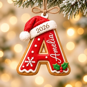 以下が含まれることがあります： ジンジャーブレッド風の文字Aのクリスマスオーナメント。赤いサンタ帽、白い縁取り、2026年の年号、白い筆記体で書かれたAmeliaの名前、白い雪の結晶、緑のヒイラギの葉と赤い実が装飾されており、茶色の紐で吊り下げられています。