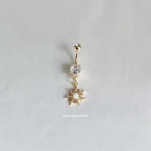 Könnte beinhalten: Goldener Bauchnabelring mit einem klaren Kristall und einem baumelnden Sonnenanhänger mit einem weißen Opal in der Mitte.