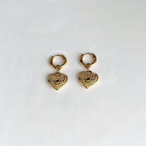 May include: Gold hoop earrings with a dangling heart charm. The heart charm has a detailed, ornate design.
