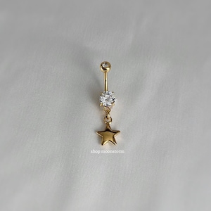May include: Gold belly button ring with a clear crystal and a dangling star charm. The charm is a gold star with a smooth, shiny finish. The text "shop moonstorm" is visible below the charm.