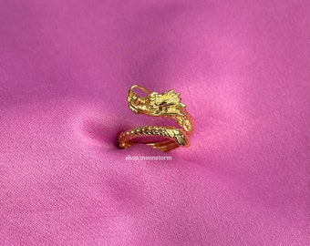 May include: A gold-colored dragon ring, coiled in a spiral, rests on a pink background. The dragon's head is detailed with scales and a small horn. The ring is a jewelry item.
