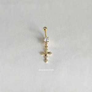 Puede incluir: Anillo de ombligo dorado con un cristal transparente y un dije de cruz con cristales transparentes.