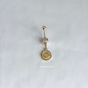 May include: Gold-toned belly button ring with a clear gem at the top and bottom. A small, round charm with a seashell design hangs from the bottom gem. The text "shop moonstorm" is at the bottom.