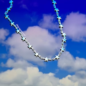 以下が含まれることがあります： 青い空と白い雲を背景に、星型のリンクが付いた銀色のチェーンネックレス。画像には「shop moonstorm」というテキストが表示されています。