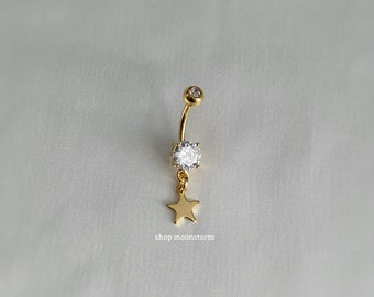 Könnte beinhalten: Goldener Bauchnabelring mit einem klaren Kristall und einem baumelnden Sternanhänger.
