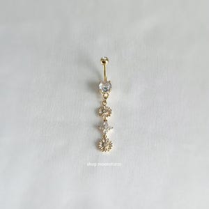 May include: Gold body jewelry with a dangling chain of three flower-shaped charms, each with clear rhinestones. The charms are connected by a small gold chain.