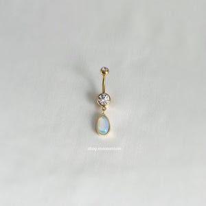 Könnte beinhalten: Goldener Bauchnabelring mit einem klaren runden Edelstein und einem tropfenförmigen Opal-Edelstein.