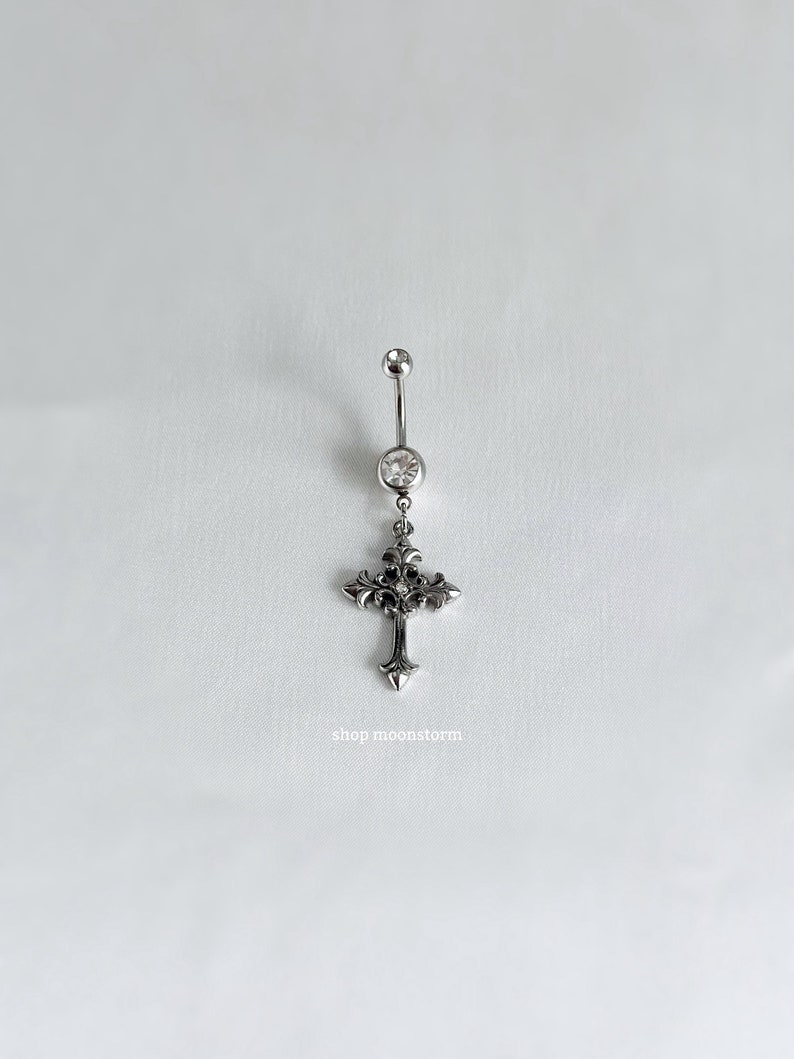 May include: Silver body piercing with a clear gem and a dangling ornate cross charm.  The charm has a detailed design with a small gem in the centre.  Shop moonstorm.