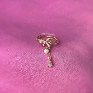 May include: Gold ring with a delicate bow design and a dangling pearl and crystal charm. The ring is set with small clear stones and is on a pink background. shop moonstorm