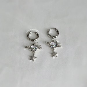 May include: A pair of silver hoop earrings with a charm of a crystal planet with a star and crescent moon dangling from it. The earrings are on a white background.