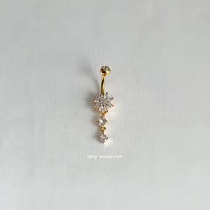 May include: Gold belly button ring with a dangling crystal flower charm. The charm has a cluster of small clear crystals.