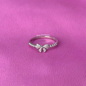 Puede incluir: Un anillo de plata con un pequeño diseño de lazo adornado con piedras de circonita cúbica transparentes. El anillo está sobre un fondo rosa.
