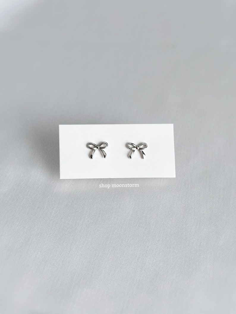 May include: A pair of silver bow-shaped stud earrings on a white card with the text "shop moonstorm".