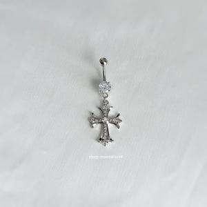 Può includere: Un piercing all'ombelico color argento con un ciondolo a forma di croce. La croce è incastonata con piccole pietre preziose trasparenti. Le sfere superiore e inferiore sono anch'esse color argento. Il testo "shop moonstorm" è in basso.