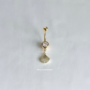 Może przedstawiać: Złoty pierścionek do pępka z przezroczystym kryształem i wiszącym urokiem w kształcie muszli. Muszla jest również ozdobiona przezroczystymi kryształami.