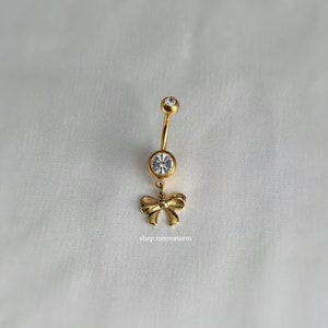 May include: Gold belly button ring with a clear crystal and a dangling bow charm. The charm is inscribed with the text "shop moonstorm".