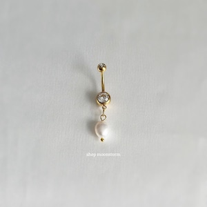 Puede incluir: Anillo de ombligo dorado con un cristal transparente y un colgante de perla blanca. El anillo está sobre un fondo blanco. shop moonstorm