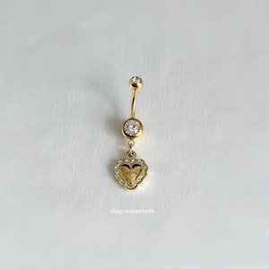 May include: Gold belly button ring with a clear gem on the top and a heart charm with a cross design on the bottom.