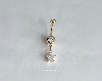 Könnte beinhalten: Goldfarbener Bauchnabelring mit einem baumelnden Sternanhänger. Der Ring hat ein gebogenes Stangendesign mit einem klaren Edelstein oben und einem größeren Edelstein in der Mitte. Der Sternanhänger besteht aus klaren, funkelnden Steinen.