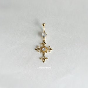 Könnte beinhalten: Goldener Körperschmuck mit einem Kreuzanhänger und klaren Strasssteinen. Das Kreuz hat ein detailliertes Design und ist mit einem kleinen Strassstein in der Mitte verziert.
