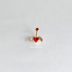 May include: Gold belly button ring with a red heart and wings. The ring is on a white background.