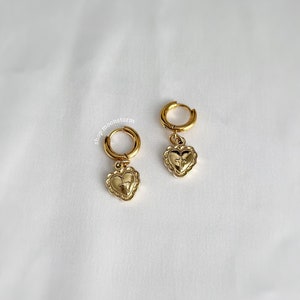 May include: Gold hoop earrings with a heart charm featuring a cross design. The earrings are on a white background.