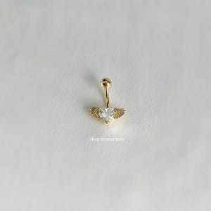 May include: A gold-colored belly button ring with a heart-shaped, clear gemstone and small wing accents. The ring has a small ball at the top and the text "shop moonstorm" is visible.