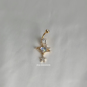 May include: Gold belly button ring with a clear iridescent crystal orb, a crescent moon, and a star with rhinestones. The ring is attached to a gold bar with a clear rhinestone on the top. The text "shop moonstorm" is visible below the ring.