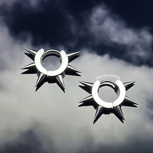 以下が含まれることがあります： エッジの周りに鋭いスパイクが付いた、銀色のフープイヤリングのペア。イヤリングは曇った空の背景に対してある。