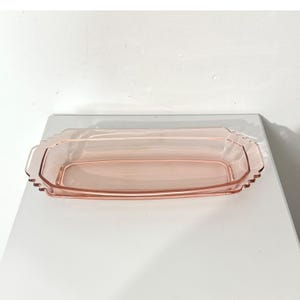 May include: A rectangular pink glass serving dish with rounded corners and a scalloped edge detail. The dish is translucent, featuring a smooth interior basin and a decorative, textured rim that extends slightly outward along the sides.